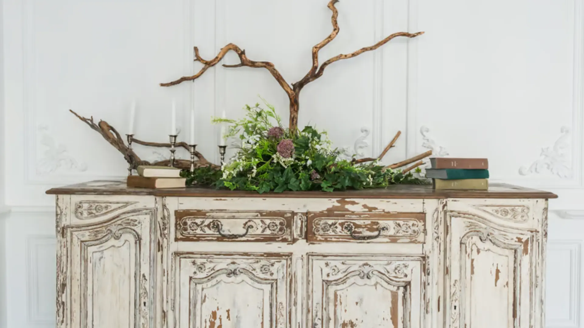 Weathered white antique sideboard styled with candlesticks, greenery and a decorative branch against a paneled wall